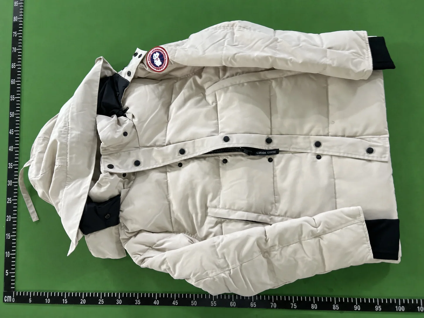 Overall front QC view of an off-white fur-trim hood parka laid against a centimetre reference board